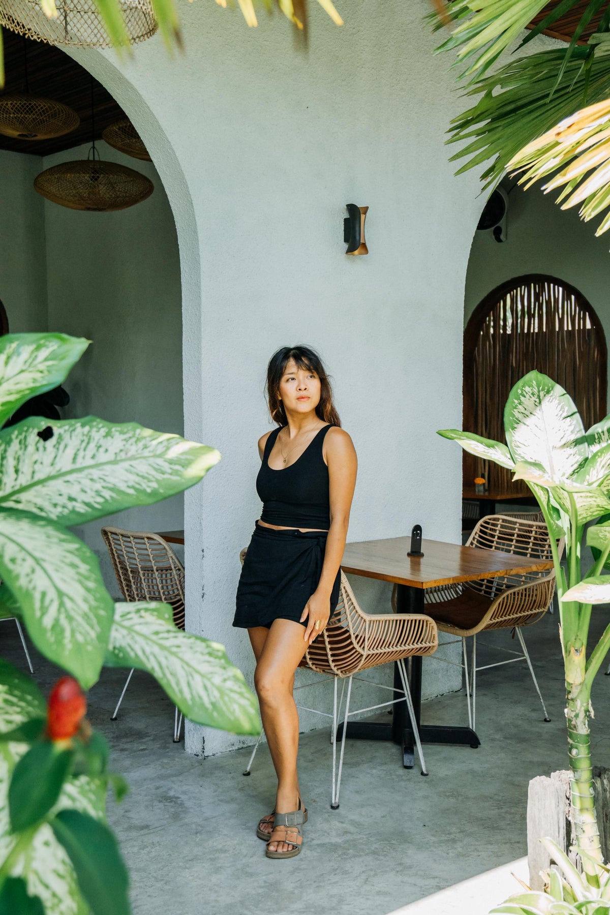A woman wearing a black sleeveless crop top and black mini skirt stands in a patio setting with woven chairs and tropical plants.