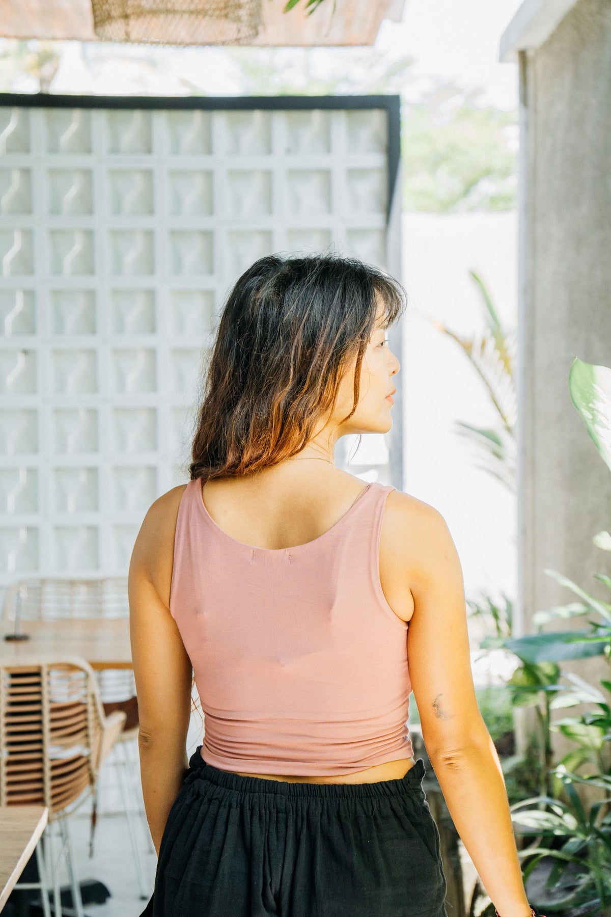 A woman viewed from behind wearing a dusty pink ribbed tank top and black bottoms, standing in a bright, modern outdoor patio setting with plants and a glass block wall.