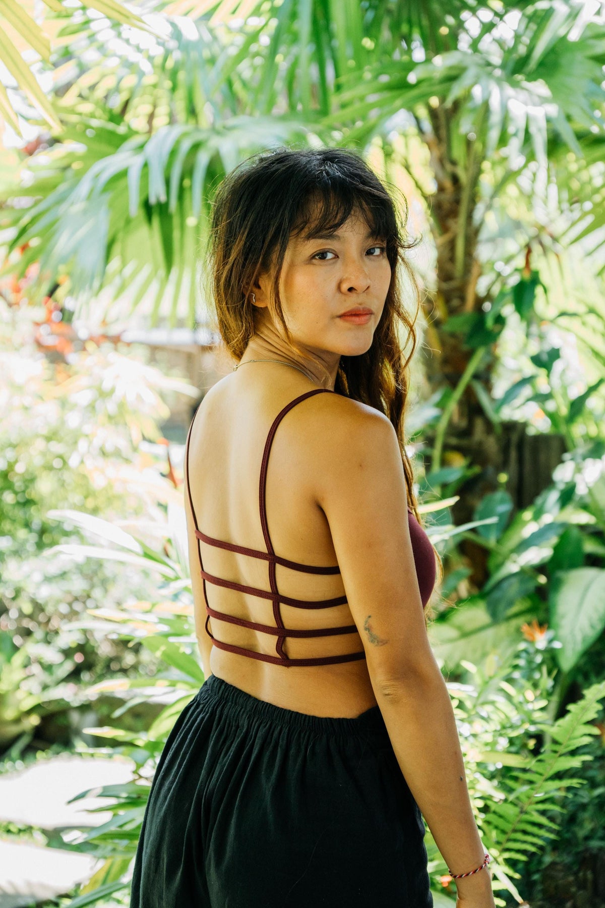 A woman wearing a dark red strappy halter top and a black skirt, photographed from the back in a lush tropical garden setting.