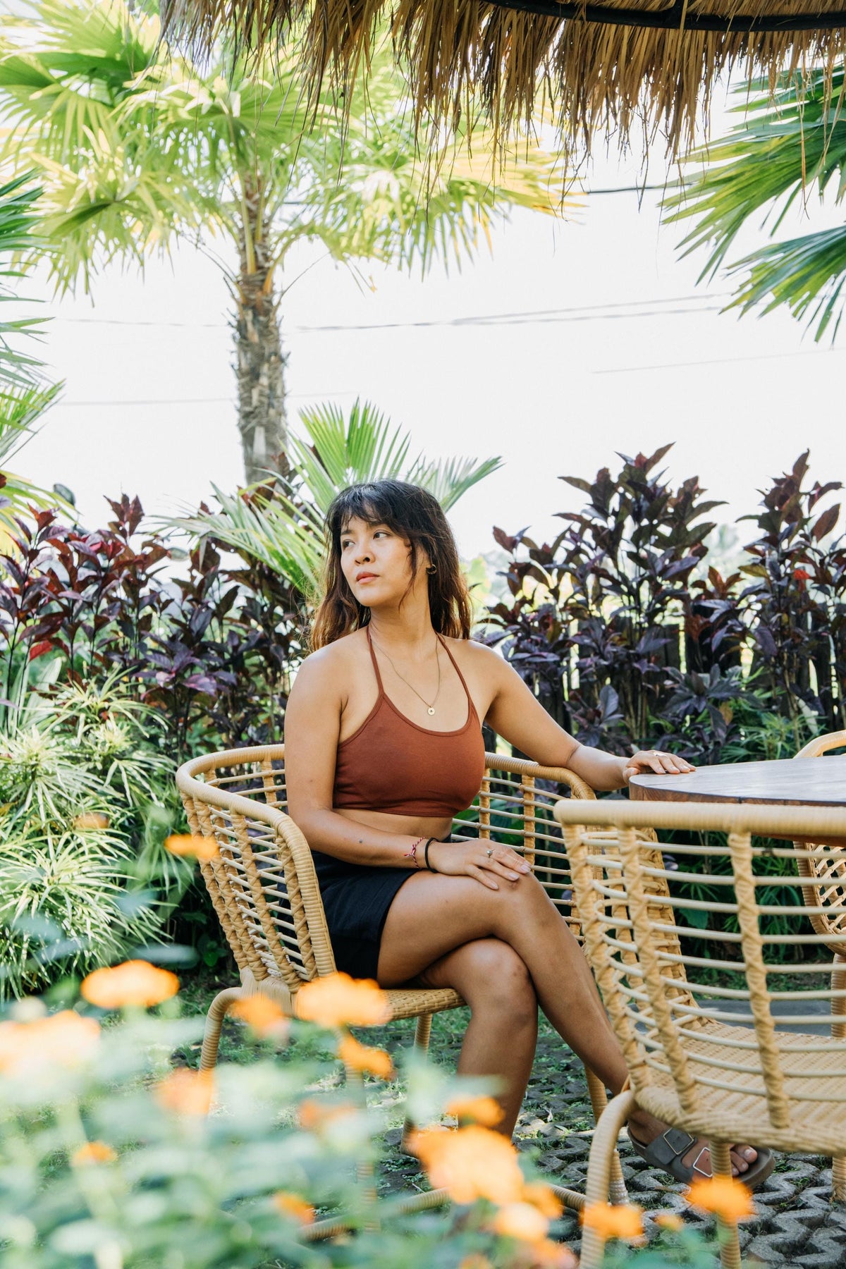 A woman sits in a woven rattan chair at an outdoor patio table, wearing a brown halter crop top and black shorts, surrounded by tropical plants and palm trees.