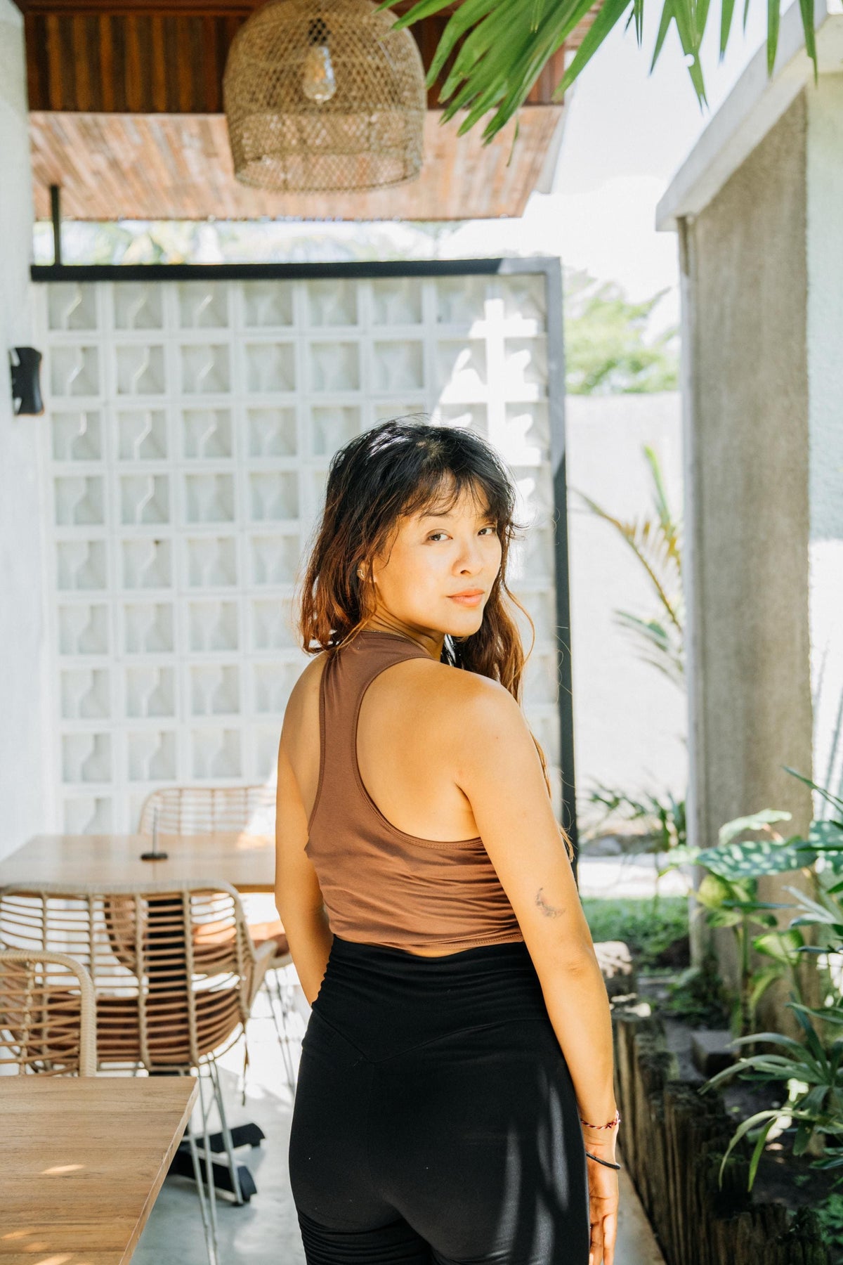 A woman wearing a brown ribbed tank top and black leggings stands in a sunlit patio setting, looking back over her shoulder. The background features a white block-patterned door, wooden furniture, and green plants.
