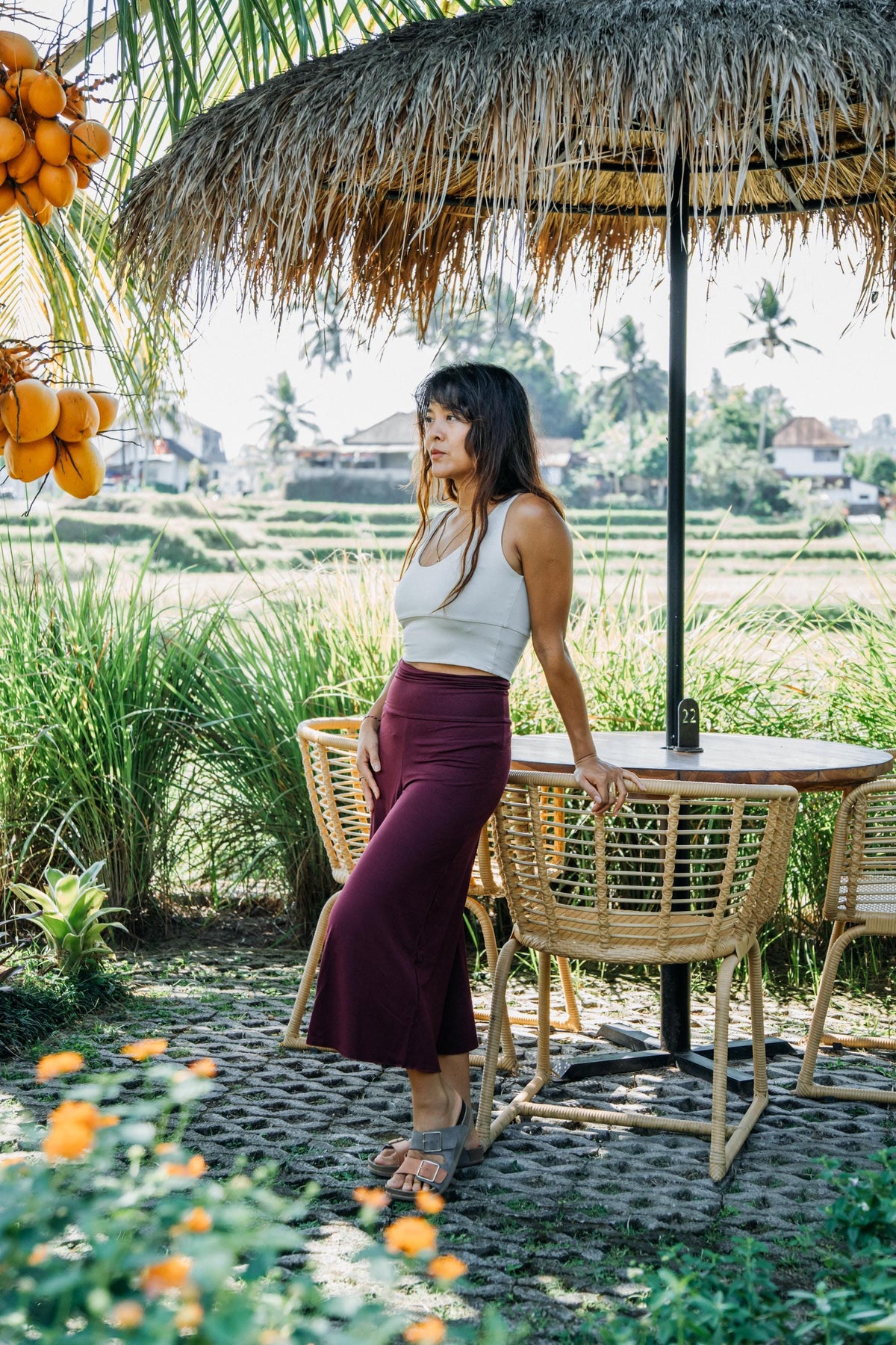 A woman stands outdoors under a thatched umbrella beside a wicker table and chairs, with a tropical rice field and palm trees in the background. She is wearing a white sleeveless top and a dark burgundy midi skirt.