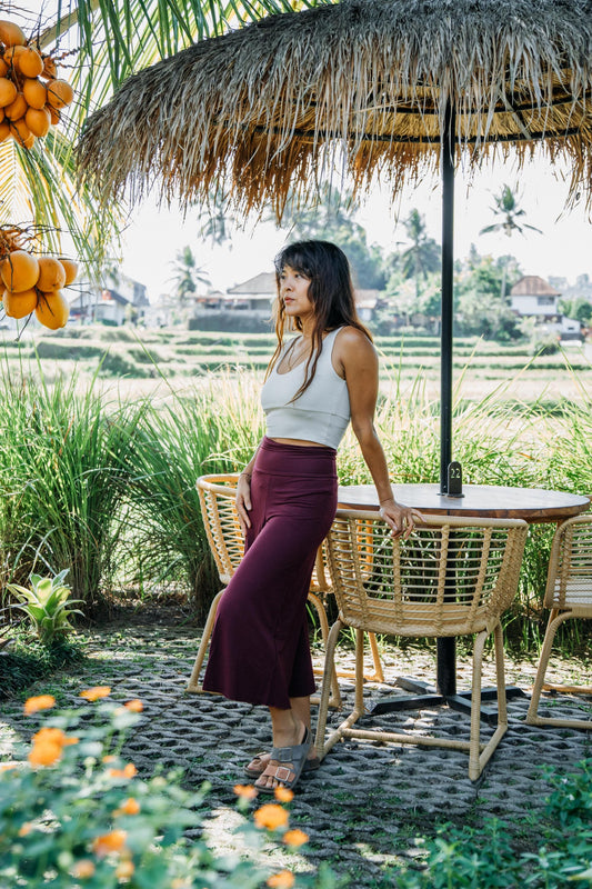 A woman stands outdoors under a thatched umbrella beside a wicker table and chairs, with a tropical rice field and palm trees in the background. She is wearing a white sleeveless top and a dark burgundy midi skirt.