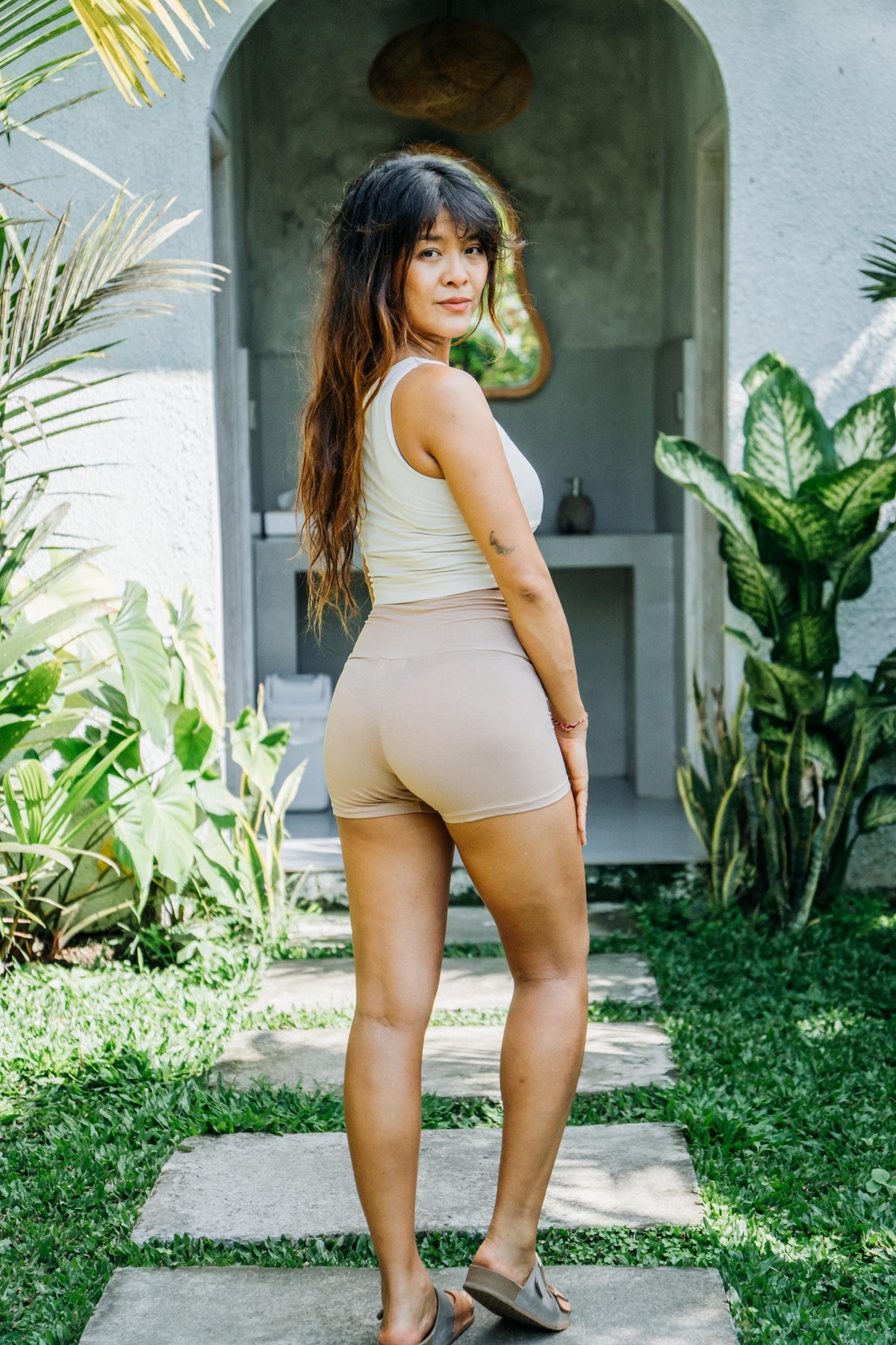 A woman wearing a beige ribbed tank top and matching high-waisted shorts stands on a stone path in a garden setting, looking back over her shoulder.