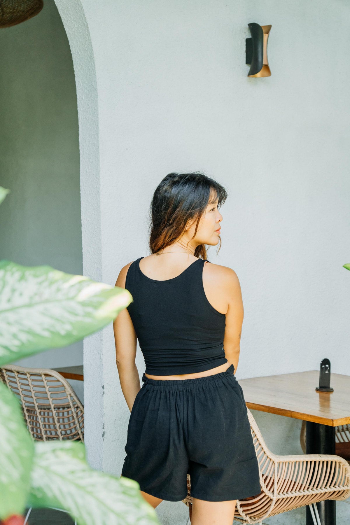 A woman wearing a black ribbed tank top and matching black shorts, photographed from behind in a bright, modern indoor setting with a light gray wall, wooden table, and woven chairs.