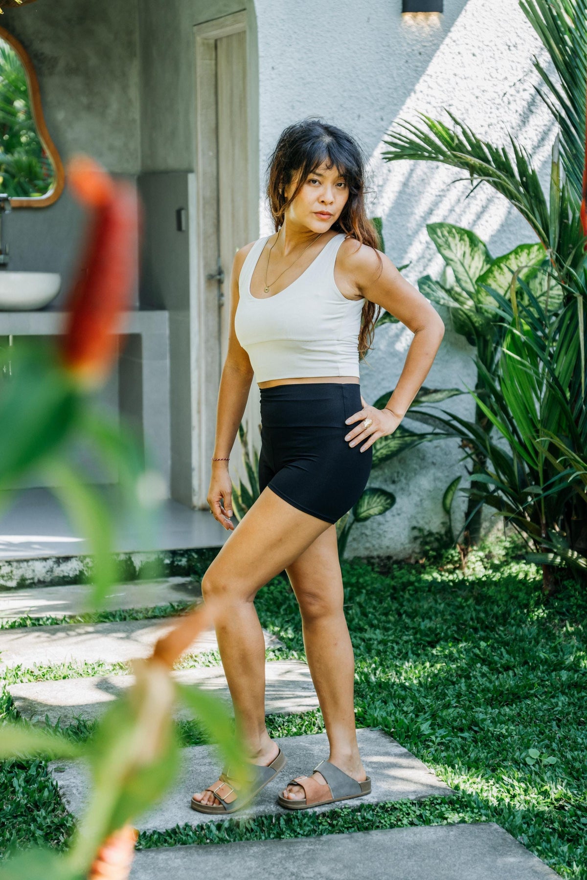 A woman posing outdoors in a white ribbed tank top and black athletic shorts, standing on a stone path with tropical plants and a modern house in the background.