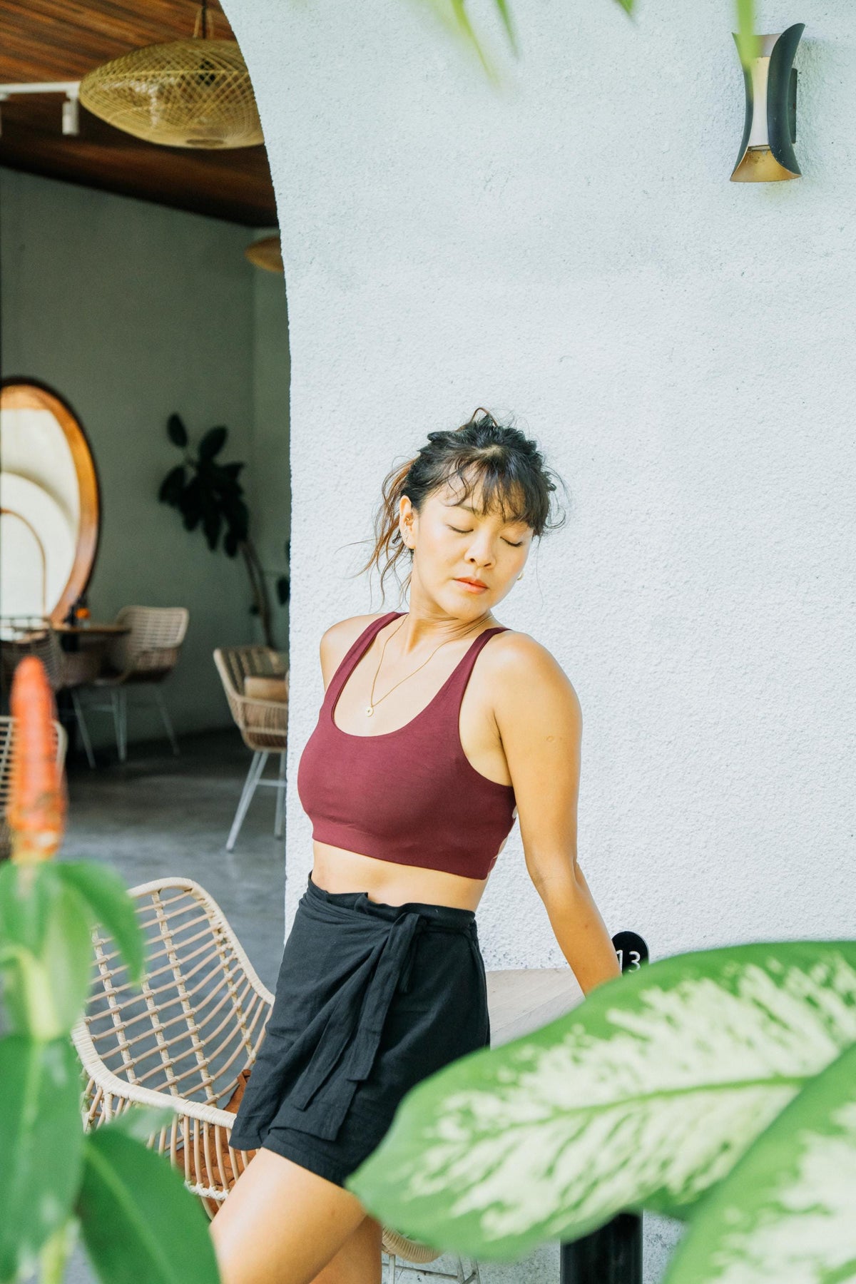 A woman wearing a maroon sports bra and black shorts posing in a modern patio setting with green plants in the foreground.