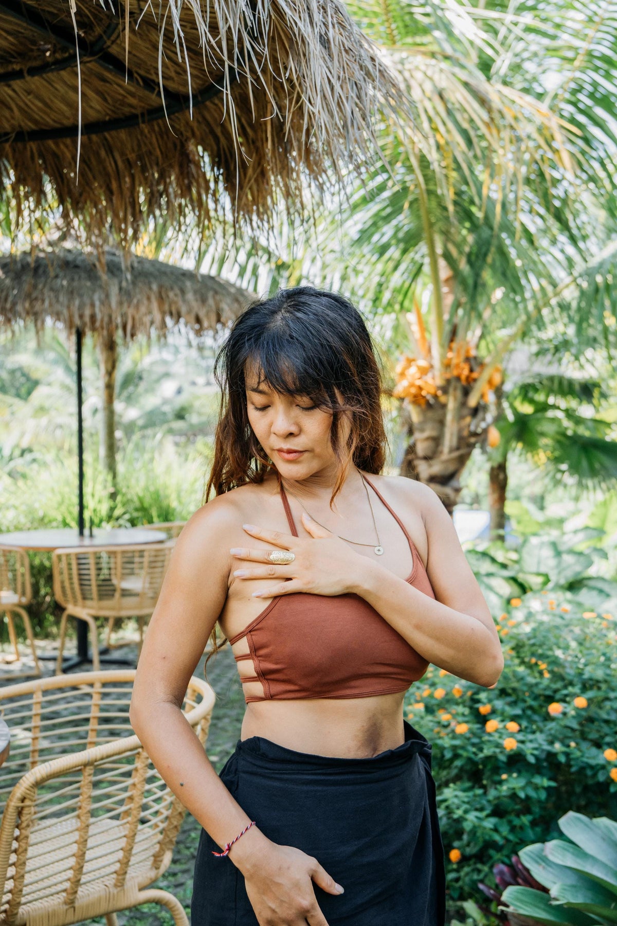 A woman wearing a rust-brown halter bikini top and a black skirt, standing outdoors in a tropical garden setting with palm trees and a thatched umbrella.